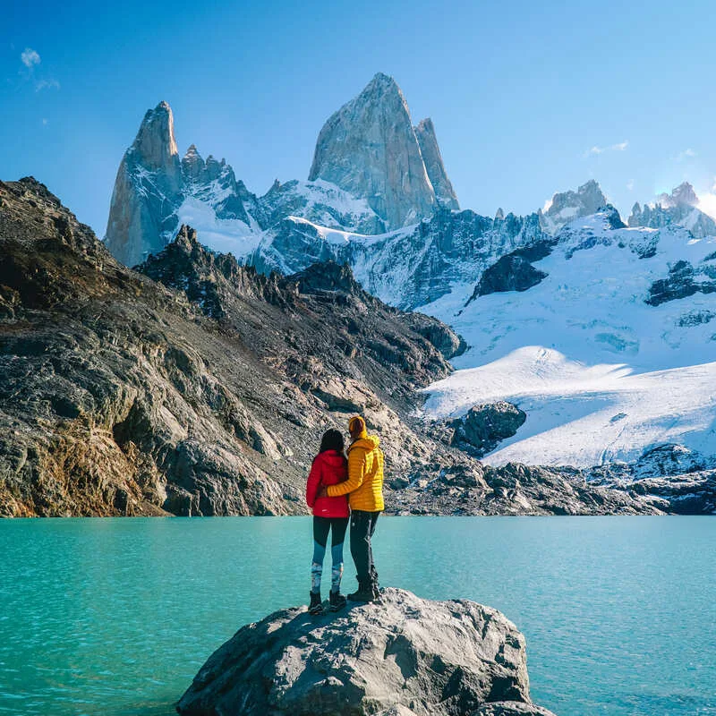 Couple Embracing As They Stand On A Rock By The Shores Of A Glacial Lake, Surrounded By Alpine, Snow Capped Peaks In Patagonia, Southern Argentina, South America