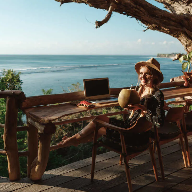 Young Female Remote Worker Enjoying A Coconut Drink While Working On Her Laptop From An Island In Indonesia, Southeast Asia, Digital Nomad Visa Concept