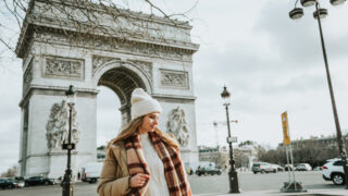 Young beautiful fashionable woman tourist in white hat and scarf on background of famous Arc de Triomphe or Triumphal arch. Winter or autumn in Europe. Paris, France.