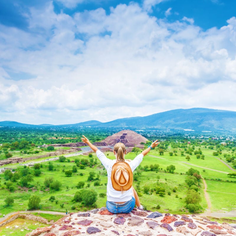 Back side of woman sitting with raised up hands on the top of mountains and enjoying view of Mexican Teotihuacan pyramid, travel and tourism concept