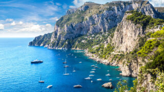Capri island in a beautiful summer day in Italy with tons of boats in the water against a high cliff.