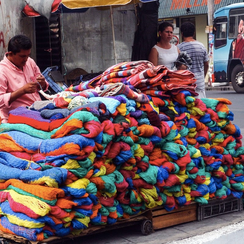 A sidewalk street vendor waits for a customer to sell one of his colorful hammocks to passersby on a busy street San Salvador.
