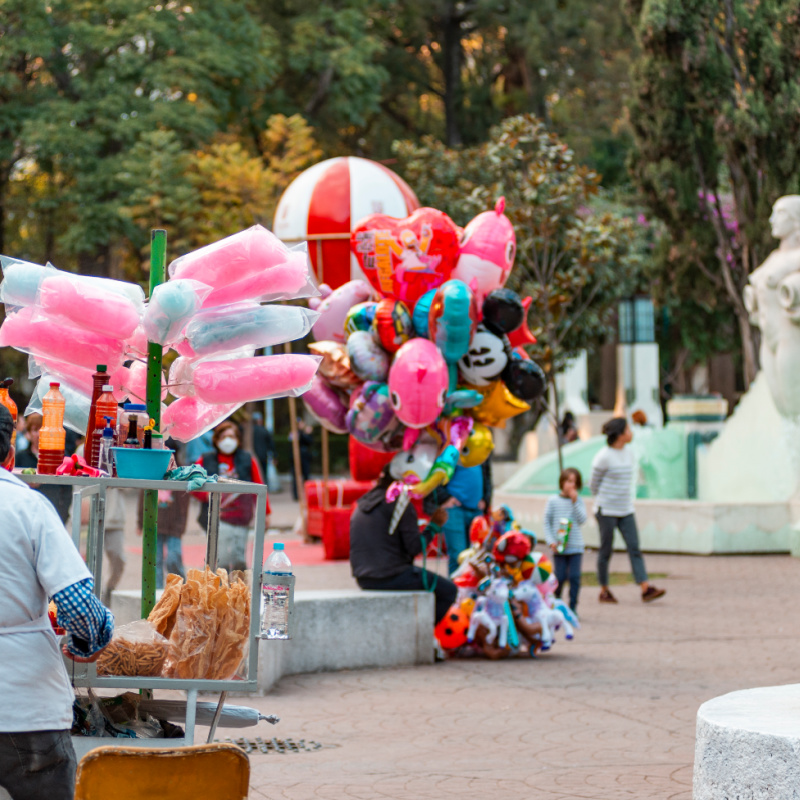 Someone sells pink and blue cotton candy, while another sells multi-colored balloons. A family walks in the background admiring the foliage. 