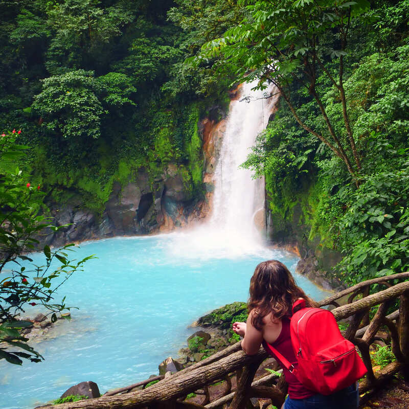 Young Female Tourist With A Backpack Watching A Waterfall In A Natural Setting, Costa Rica, Central America