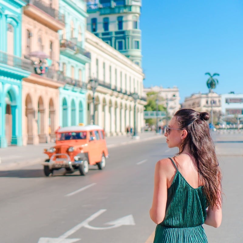 Young Woman In A Summery Dress Wandering The Pastel-Colored Historic Streets Of Havana, Cuba As An Orange Vintage Car Approaches, Cuba, Latin America