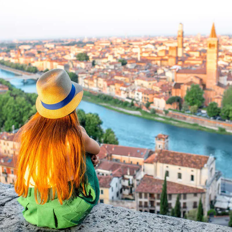 A Young Woman Wearing A Straw Hat As She Sits On A Wall Atop A Viewpoint With A Panorama Of The City Of Verona, In Northern Italy, Europe