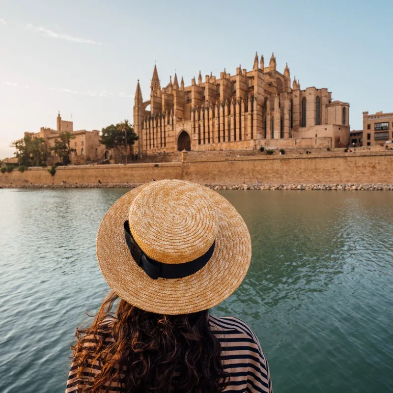 Female Tourist With A Straw Hat Watching The Sun Set Over The Cathedral In Palma De Mallorca, Balearic Islands, Spain