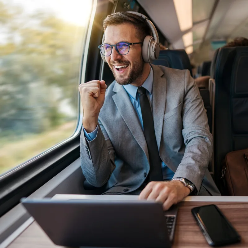 a man in a suit looks excited as he sits on a train with headphones on and a computer open