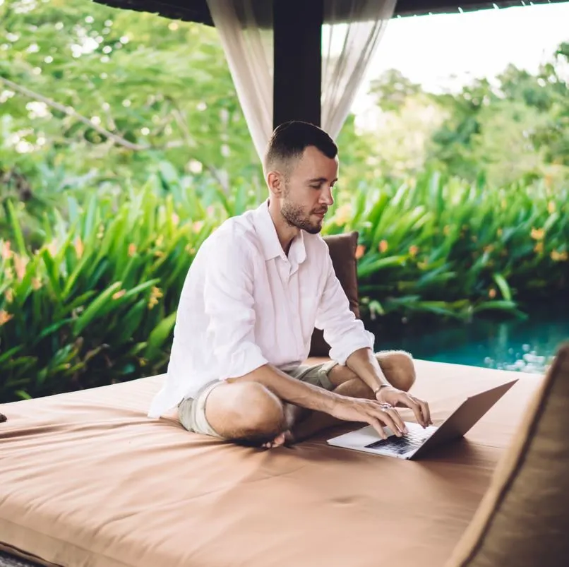 Man working on his laptop on a day bed next to a pool