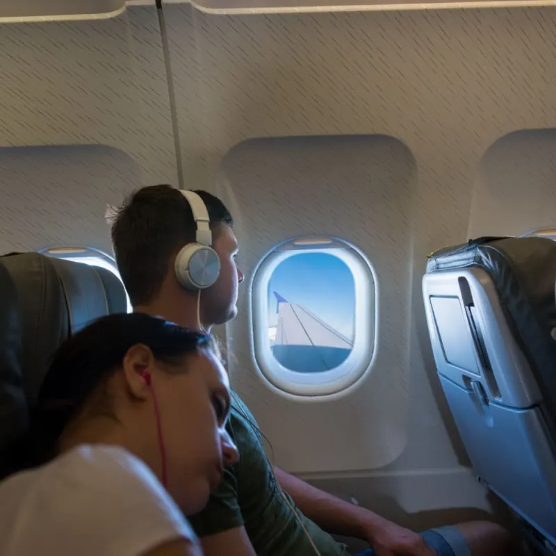 Female Passenger Sleeping On A Plane As Her Partners Gazes Out The Window Listening To Music In His Headphones, Unspecified Airline
