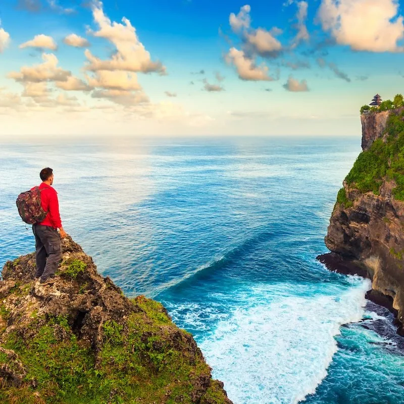 Man backpacker on the top of the hill enjoy scenery view hindu temple Uluwatu temple in Bali Indonesia on background amazing nature at sunrise  / Man hiker on a top of a mountain / Bali, Indonesia