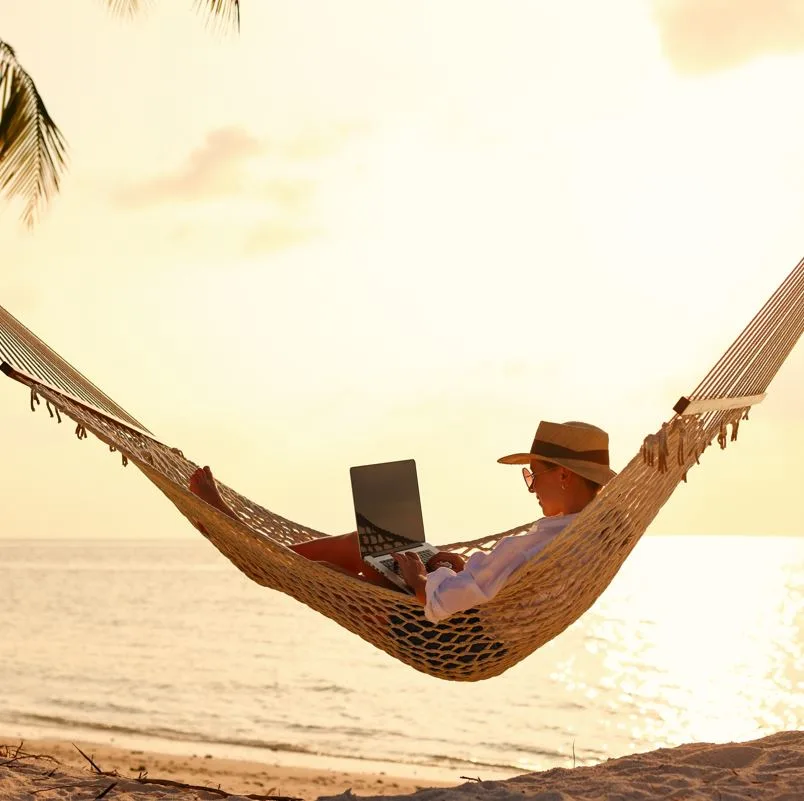 Woman working on laptop in a hammock over the sand next to the ocean