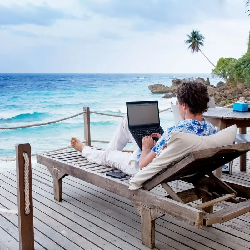 Young Male In A Floral Shirt, Summery Outfit Working From His Computer In A Beach Setting, Digital Nomad Concept