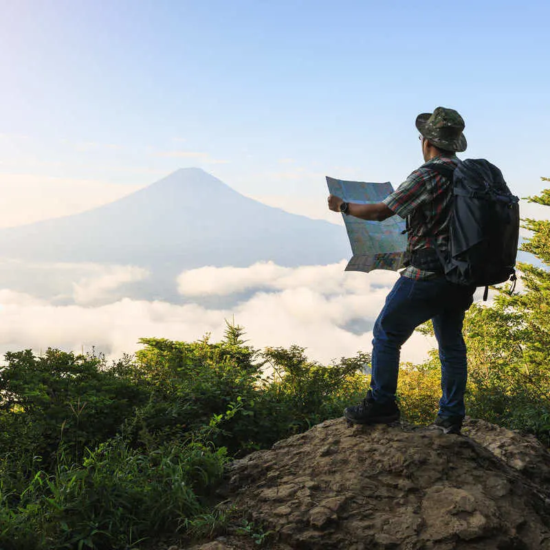 Young Male Tourist Holding Up A Map As He Gazes At A Mountain Range In The Distance, Nature Travel, Japan, Southeast Asia